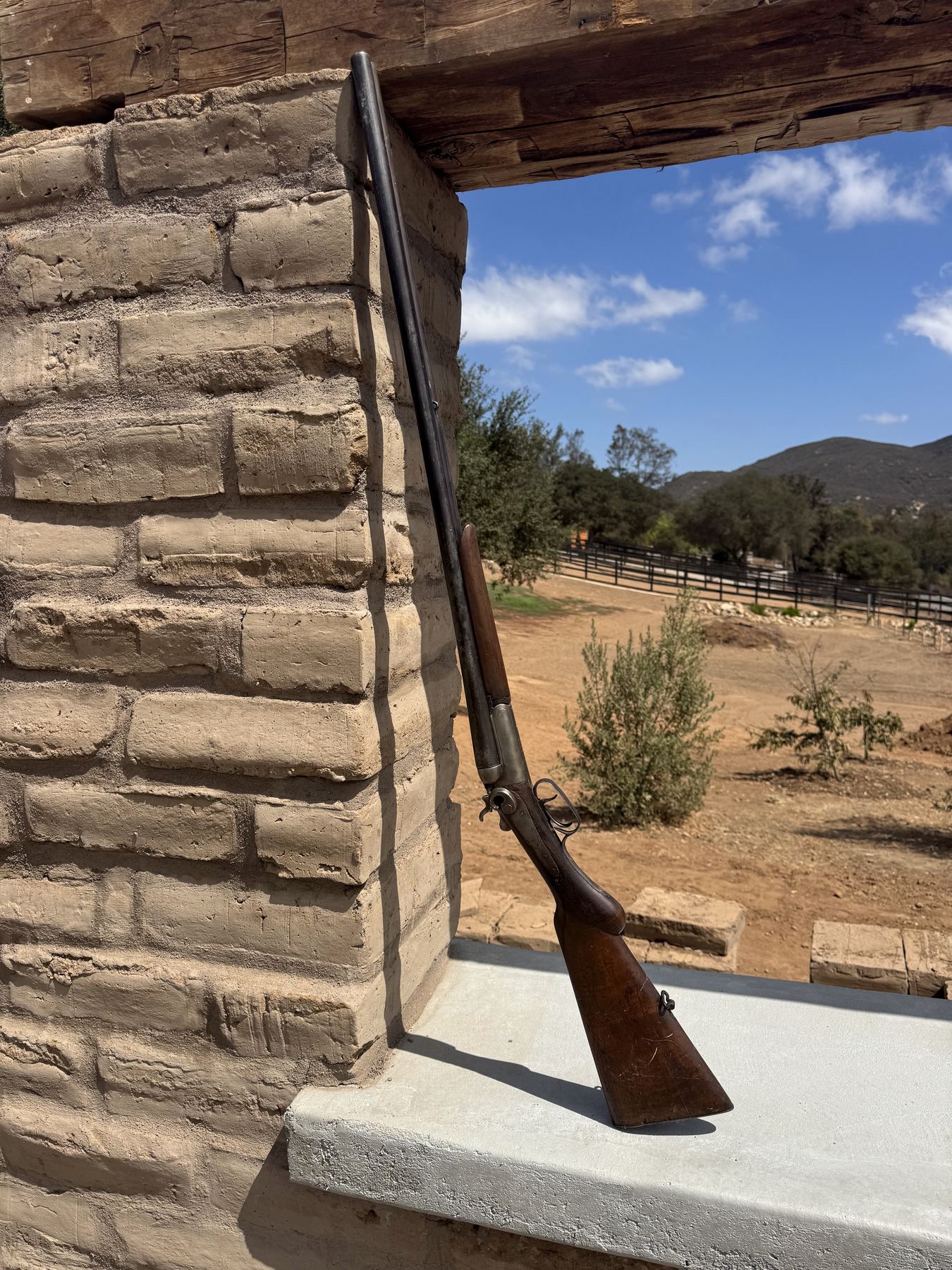A double-barreled shotgun leaning on a reconstructed adobe wall in an open window, with the Elfin Forest hills beyond.