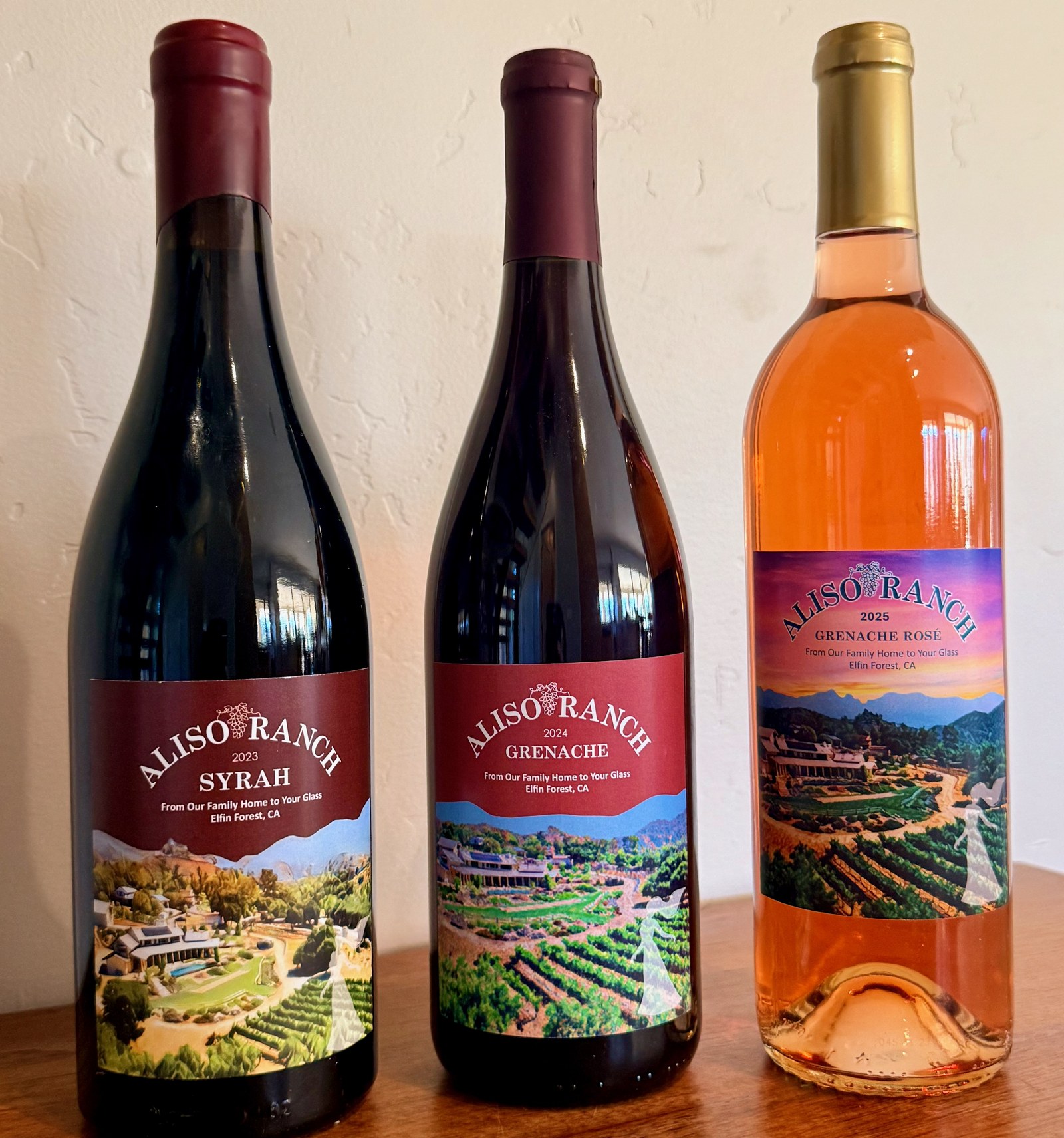 Three Aliso Ranch bottles on a wood table: 2023 Syrah, 2024 Grenache, and 2025 Grenache Rosé